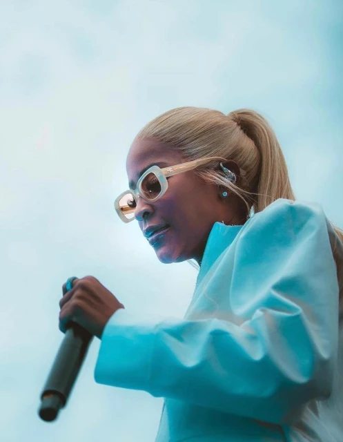 Woman sings into a microphone, wearing sunglasses and a white jacket with blonde hair in a ponytail against a light blue background.
