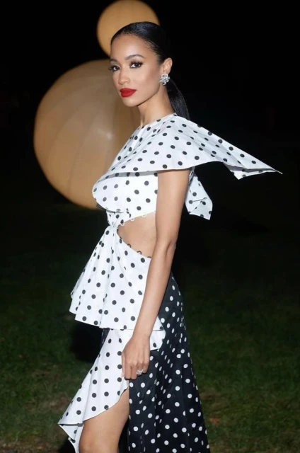 Elegant woman poses in a black and white polka dot dress with side cut-out, red lipstick, and diamond earrings against a dark backdrop.