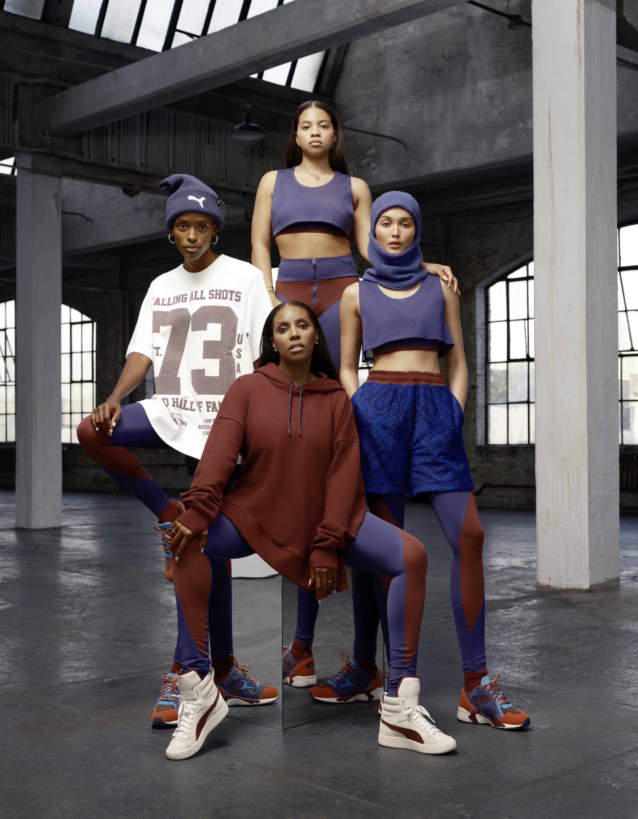 Four women model Puma athletic wear in a studio setting with exposed brick and natural light, showcasing matching outfits and sneakers.