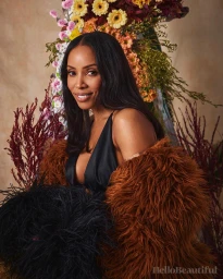 Portrait of a woman with long dark hair wearing a black top and a textured brown faux fur coat against floral arrangements