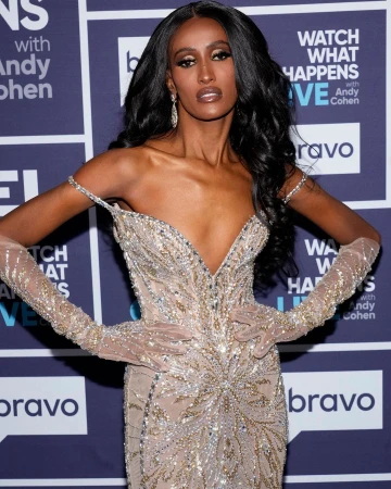 Woman with long dark hair wearing a sparkly beaded gown and sheer gloves posing against a purple branded backdrop.