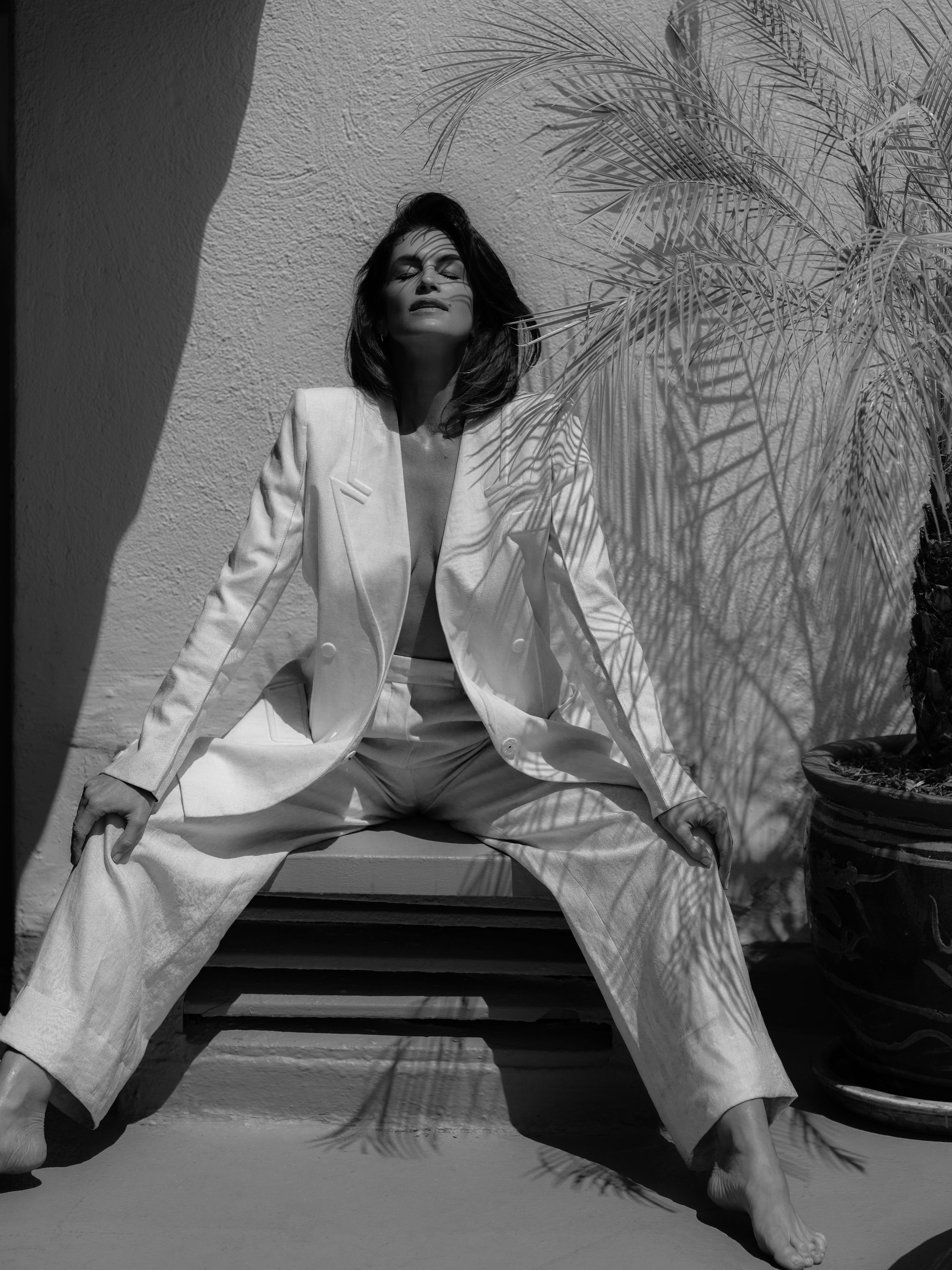 A woman in a white suit sits in sunlight with a palm tree casting shadows. Fashionable and elegant, the image is in black and white.