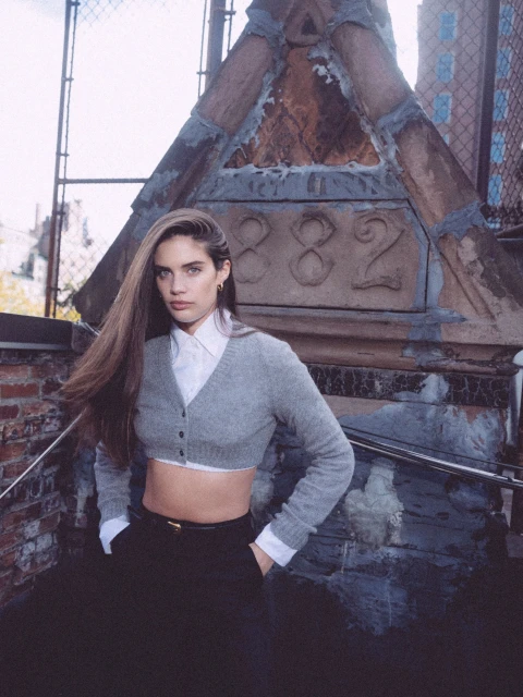 Attractive young woman with long brown hair poses in front of architectural stone steps in a gray crop top and black pants, outdoors.