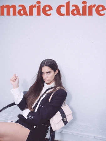 Model seated in a chair wearing a black and white trimmed jacket with a quilted shoulder bag against a plain wall.