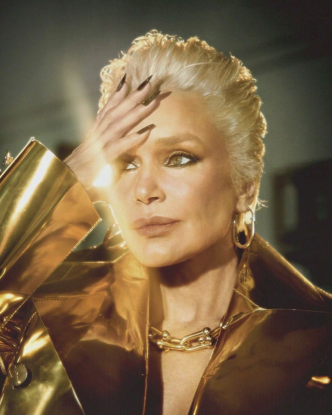 Close-up portrait of an older woman with platinum blonde hair wearing a gold jacket and gold jewelry, her hand raised to her forehead.