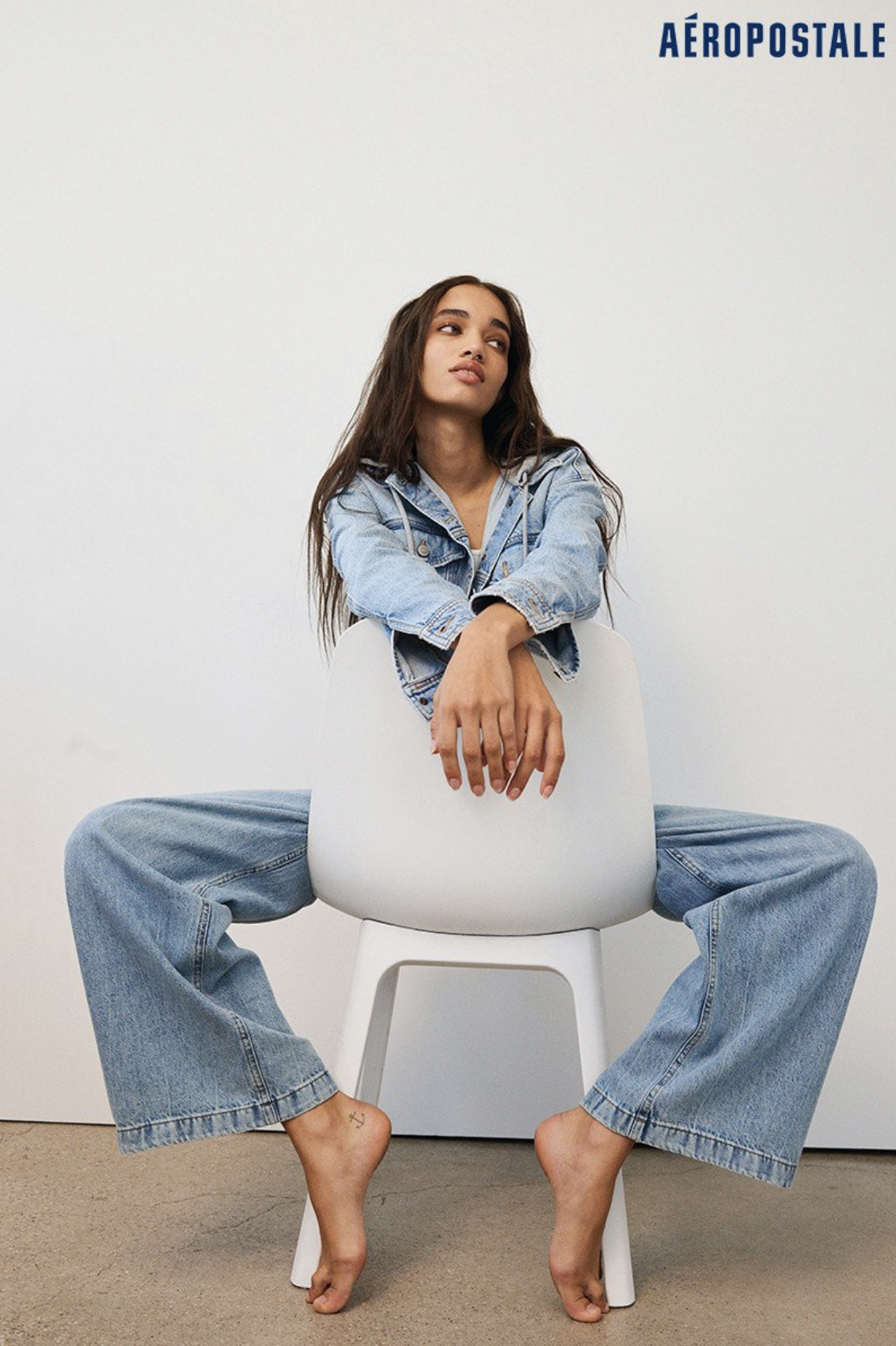 A young woman sits on a white chair against a white wall, wearing a denim jacket and wide-leg jeans by Aeropostale; looking off-camera.