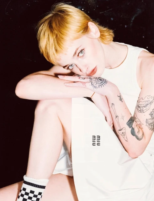 Young woman with tattoos and blonde hair rests her head on her hands while posing in front of a dark background and wearing brand-name clothing.