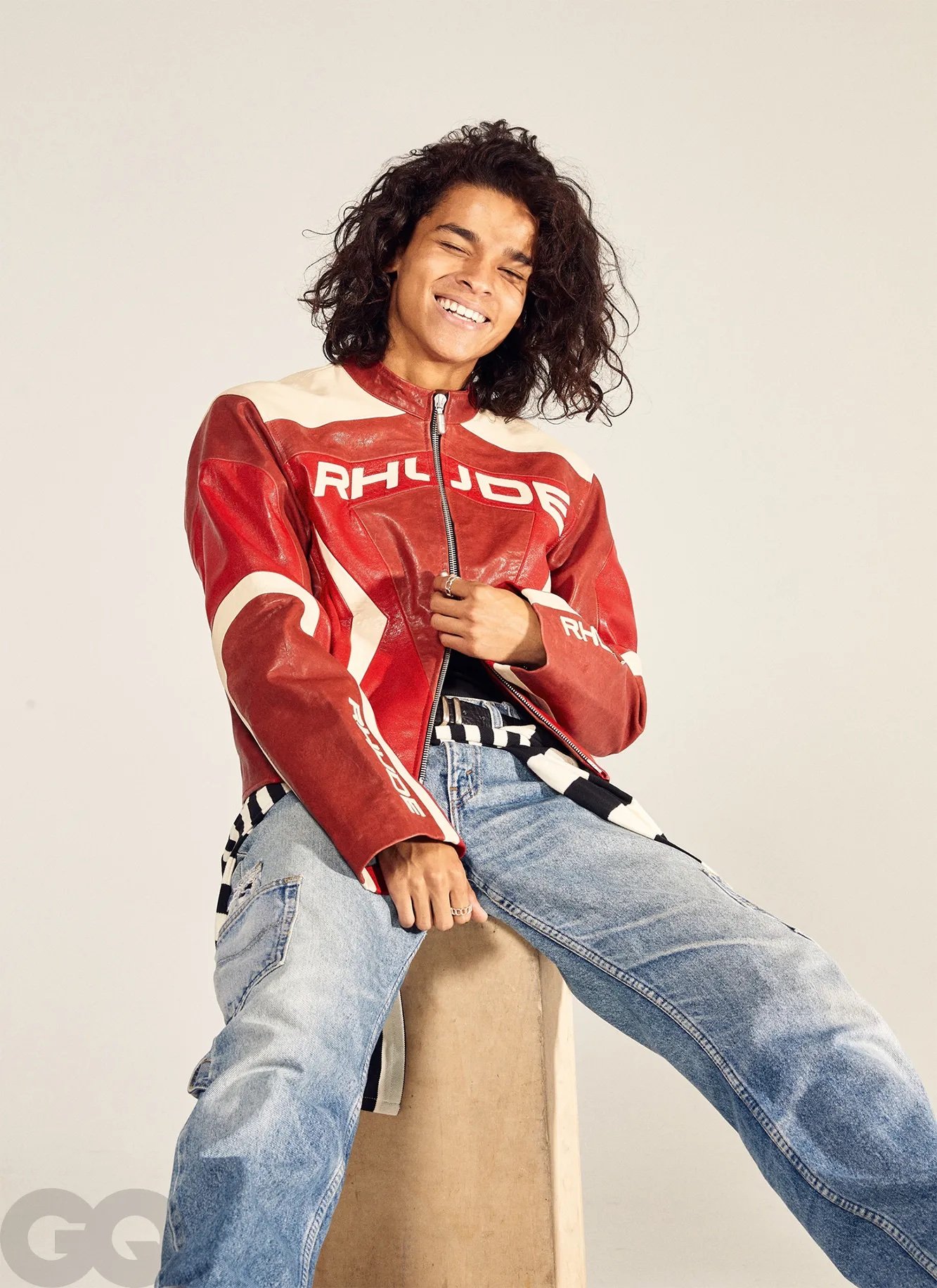Smiling male model with long curly hair, wearing a red and white leather jacket and blue jeans, sitting on a stool in a studio.
