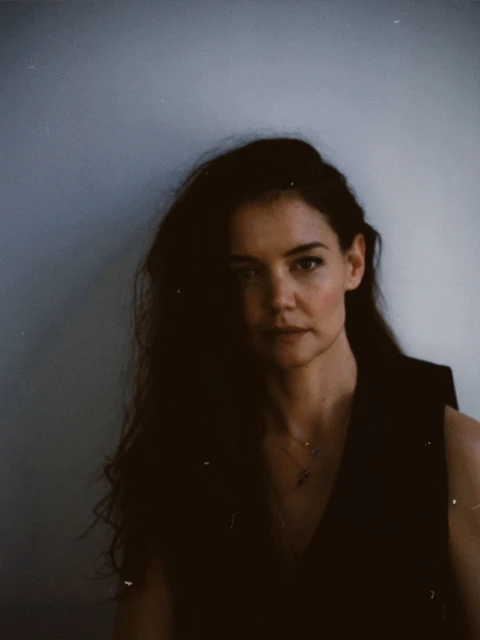 Portrait of a brunette woman with wavy hair wearing a dark top, standing against a light gray wall in a photo studio.