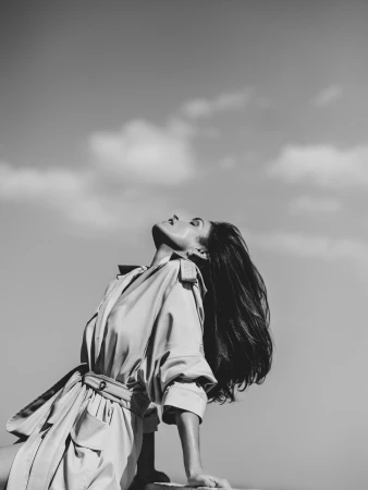 Black and white portrait of a woman with long dark hair wearing a belted trench coat against a cloudy sky backdrop.