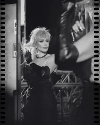 Black and white portrait of a blonde woman adjusting her hair in a mirror, wearing long dark gloves and a necklace.