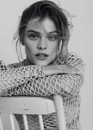 Portrait of a woman with textured hair leaning on a wooden chair wearing an open weave knit top against a plain light background