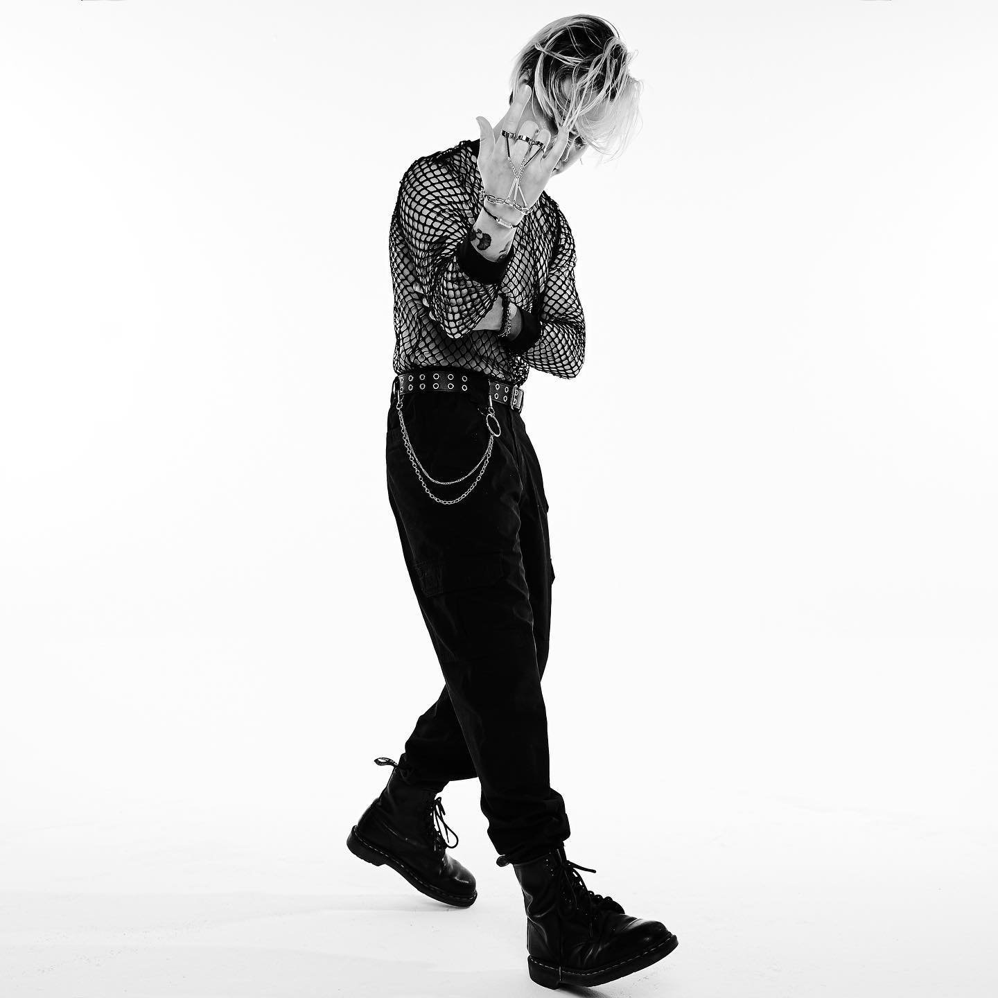 Full-length black and white studio shot of a young adult man wearing a mesh top, black pants, and boots. He has blonde hair and multiple rings.