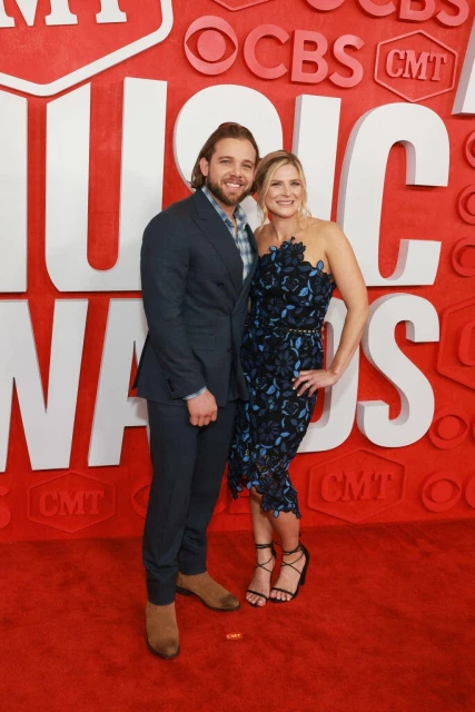 Man and woman pose on a red carpet in evening wear with a large patterned background for a music awards event.