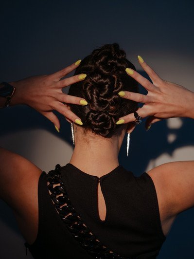 Woman with an updo hairstyle and yellow manicure displays hands behind her head. The black dress and earrings complete the look.