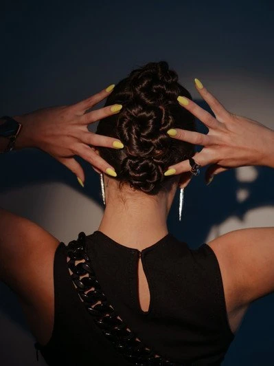 Woman with an updo hairstyle and yellow manicure displays hands behind her head. The black dress and earrings complete the look.