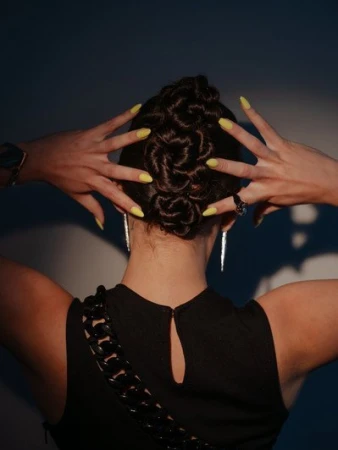 Woman with an updo hairstyle and yellow manicure displays hands behind her head. The black dress and earrings complete the look.