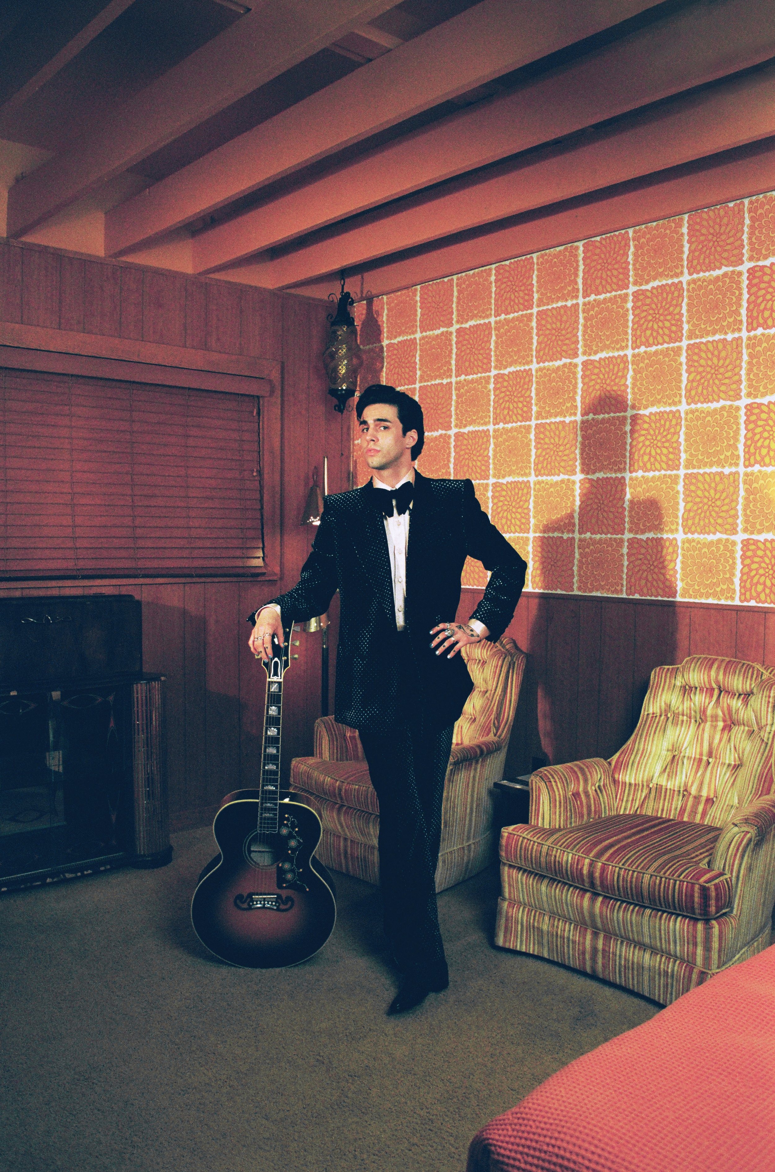 Full shot of a dark-haired man in a black tuxedo posing with an acoustic guitar in a room with orange patterned wallpaper and wood paneling.