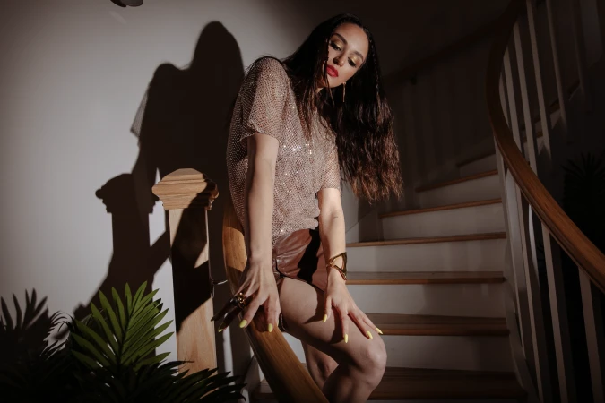 Brunette woman in a gold mesh top and brown shorts poses on wooden stairs in front of a wall with shadows and a tropical plant.