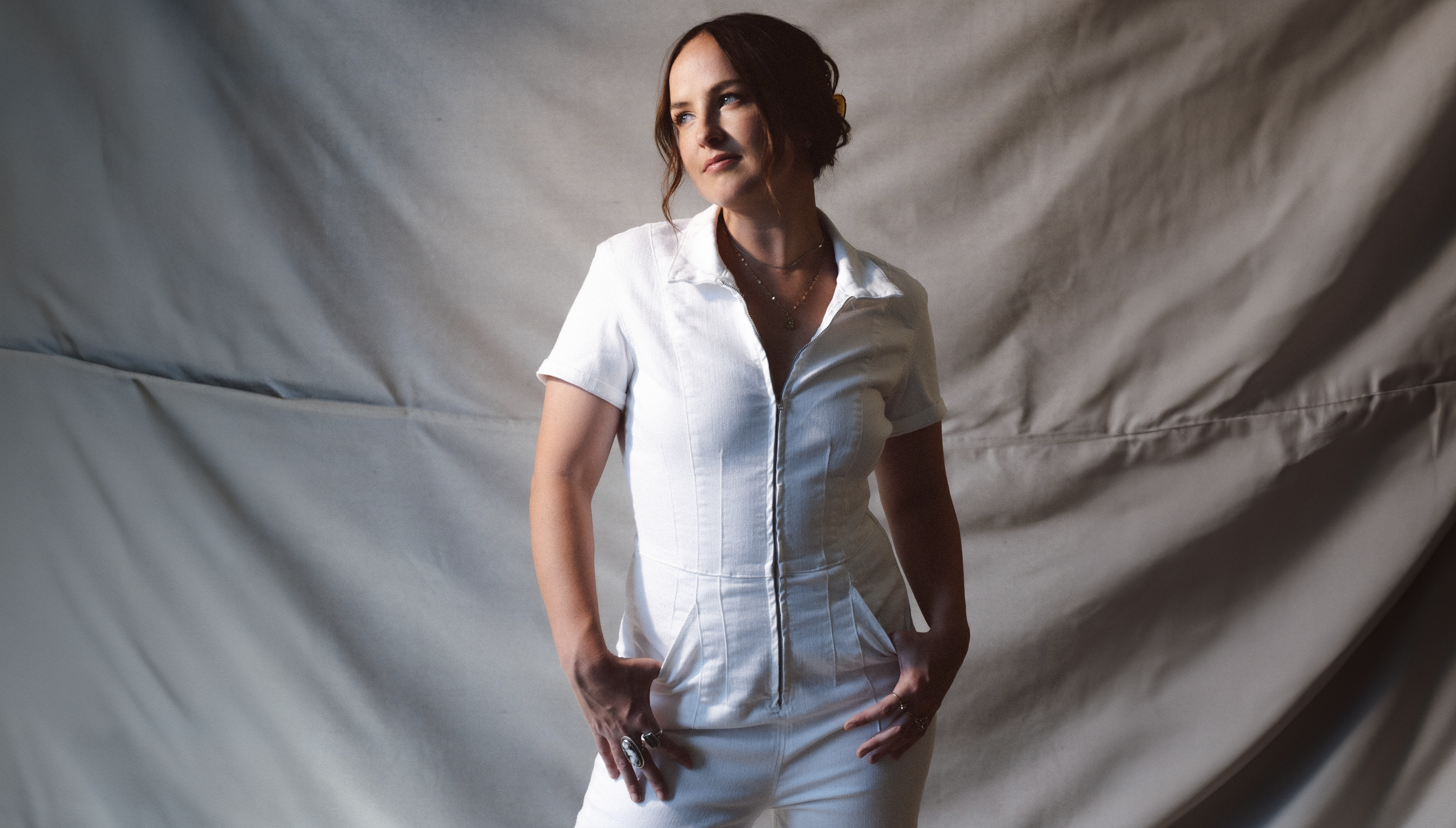 Woman in white jumpsuit poses in front of draped fabric. Fashion shot featuring adult female in studio-like environment with neutral backdrop.