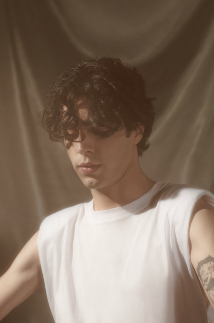 Close-up studio portrait of a young adult male with dark curly hair, wearing a white sleeveless shirt against a draped neutral background.
