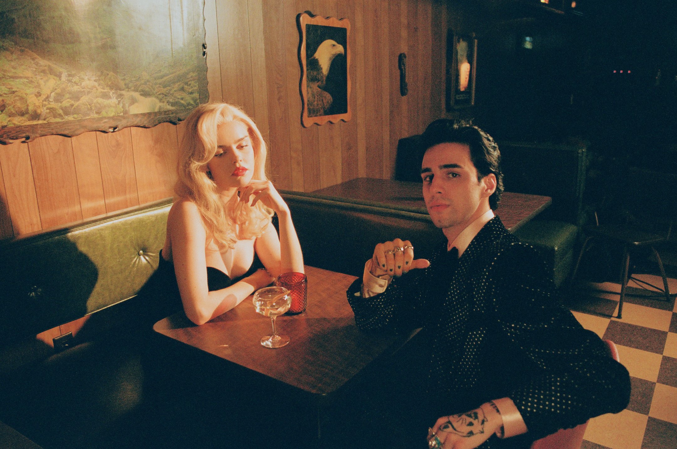 A couple sits in a vintage-style restaurant booth at a table with a champagne glass, featuring a man in a spotted suit and a woman with blonde hair.