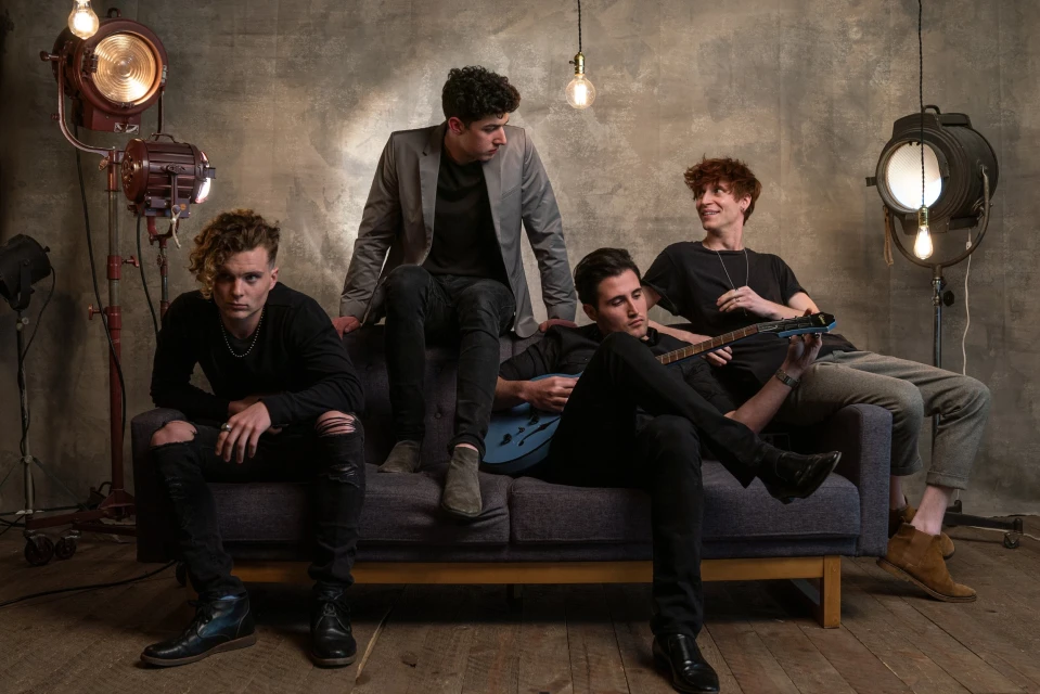 Four young adult men are posing on a couch in a studio setting with stage lighting, one playing an electric guitar.