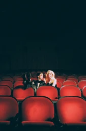 Two people seated in an empty vintage movie theater with red velvet seats, with make-up by Risa Rodriguez.