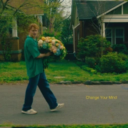 A young man with curly red hair carrying a large bouquet of flowers while walking along a suburban street.