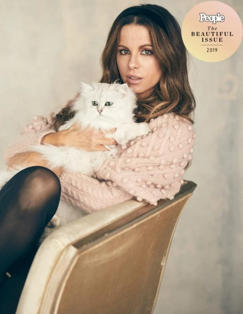 Kate Beckinsale poses with her white long-haired cat while sitting on a gold chair in a photo studio with a textured background.