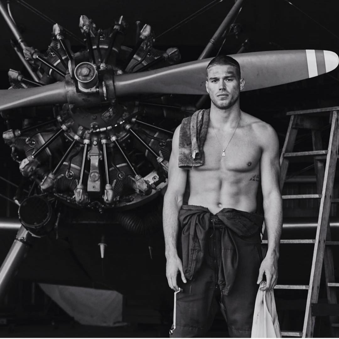 Monochrome photo of a muscular shirtless man in overalls standing in front of an airplane engine, with a towel draped over his shoulder.