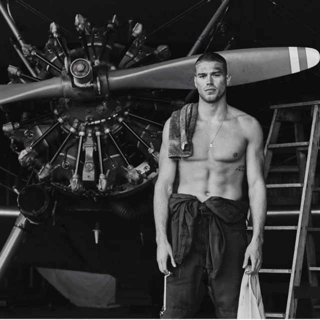 Monochrome photo of a muscular shirtless man in overalls standing in front of an airplane engine, with a towel draped over his shoulder.