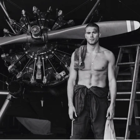 Monochrome photo of a muscular shirtless man in overalls standing in front of an airplane engine, with a towel draped over his shoulder.