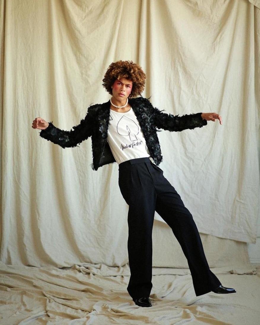 Fashionable adult posing in black jacket and pants in a studio, set against a neutral backdrop, exudes a blend of confidence and style.