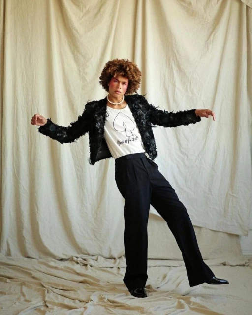 Fashionable adult posing in black jacket and pants in a studio, set against a neutral backdrop, exudes a blend of confidence and style.
