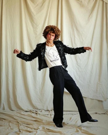 Fashionable adult posing in black jacket and pants in a studio, set against a neutral backdrop, exudes a blend of confidence and style.