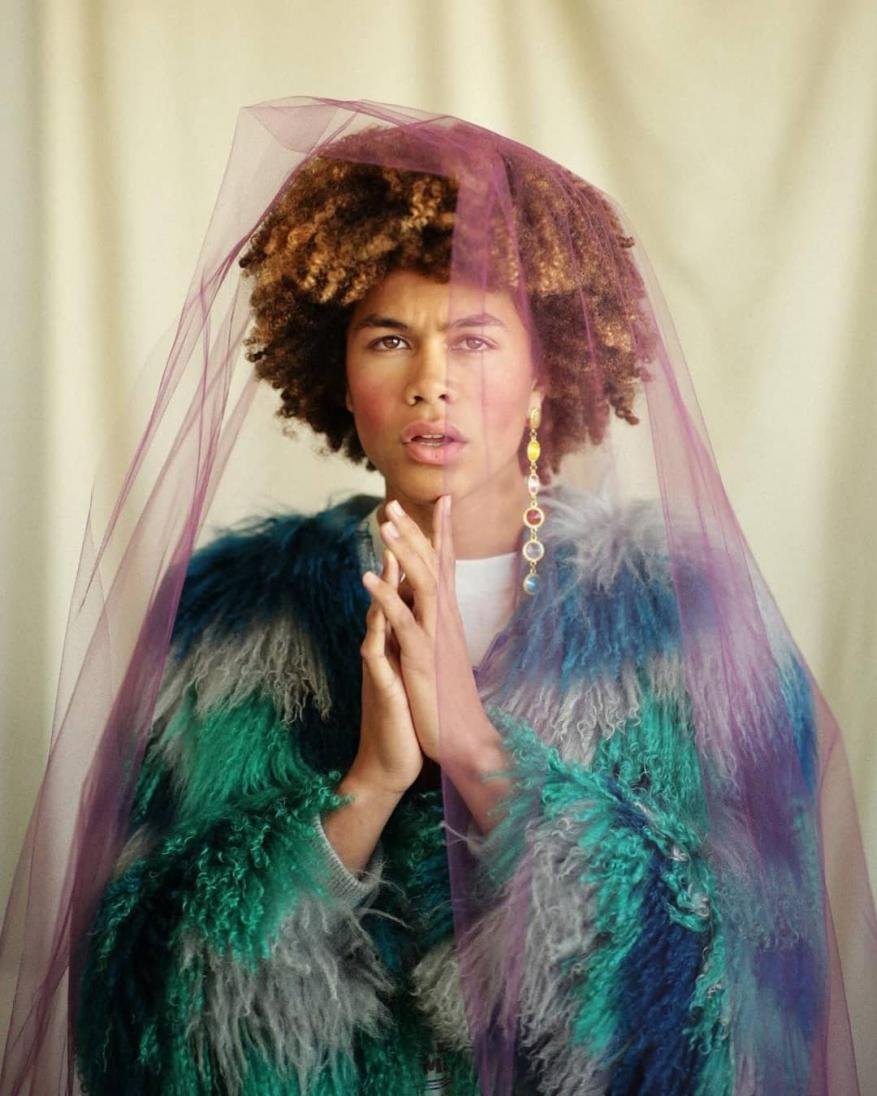 Portrait of person with curly hair wearing a blue faux fur coat and purple tulle veil with crystal drop earrings