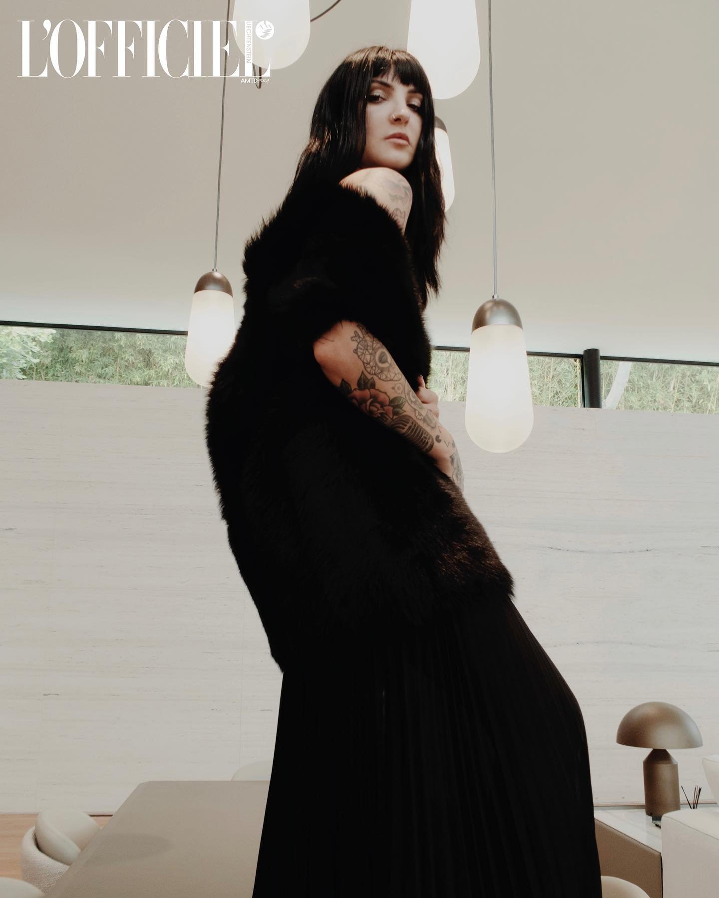 A fashionable brunette woman in a black dress poses indoors under modern pendant lights in front of a neutral background.