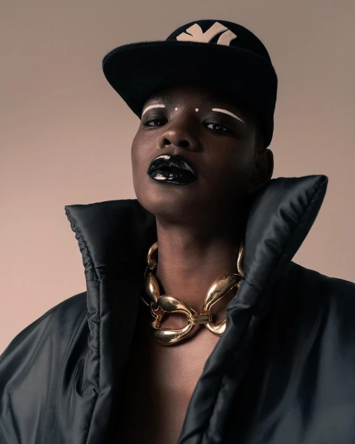 Fashionable portrait of a woman with dark skin wearing glossy black lipstick, a necklace, and a baseball cap on a beige background.
