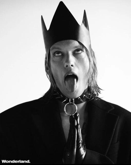 Black and white portrait of a person wearing a crown and spiked choker, sticking their tongue out against a bright background.