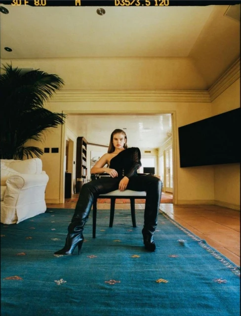 Fashion shot of a woman in a black one-shoulder top and leather pants and boots, sitting on a chair in an indoor setting.