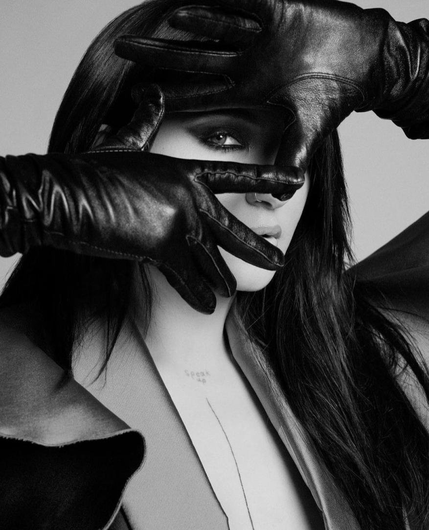 Monochrome portrait of a woman with long, dark hair covering her face with black leather gloves, wearing a coat against a grey backdrop.