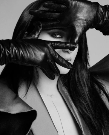Monochrome portrait of a woman with long, dark hair covering her face with black leather gloves, wearing a coat against a grey backdrop.