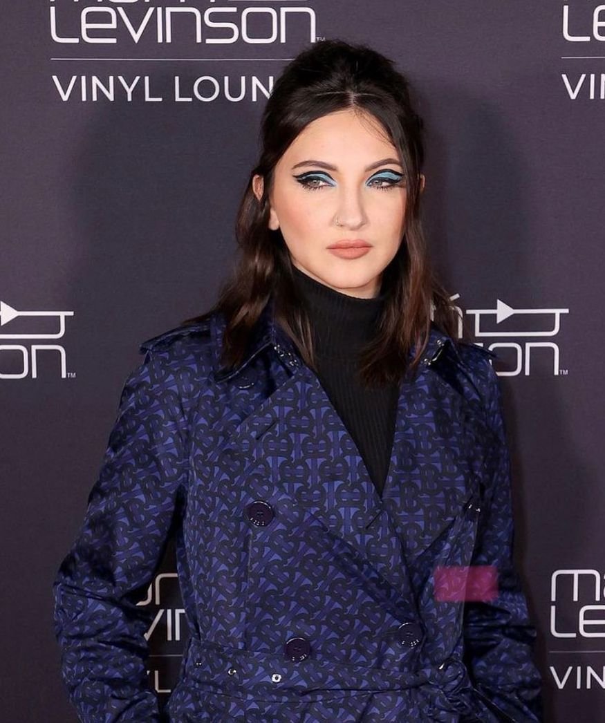 Brunette woman in a patterned trench coat poses in front of a sign for Martin Levinson's Vinyl Lounge. She has blue eyeshadow.