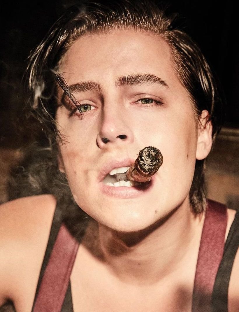 Headshot of a young adult man with dark hair smoking a cigar, wearing suspenders against a dark background in a medium shot.