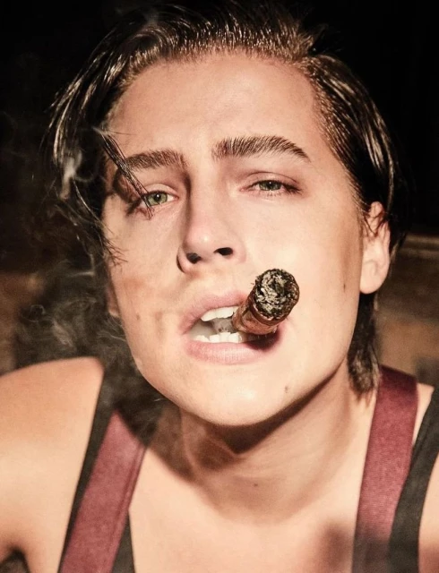 Headshot of a young adult man with dark hair smoking a cigar, wearing suspenders against a dark background in a medium shot.