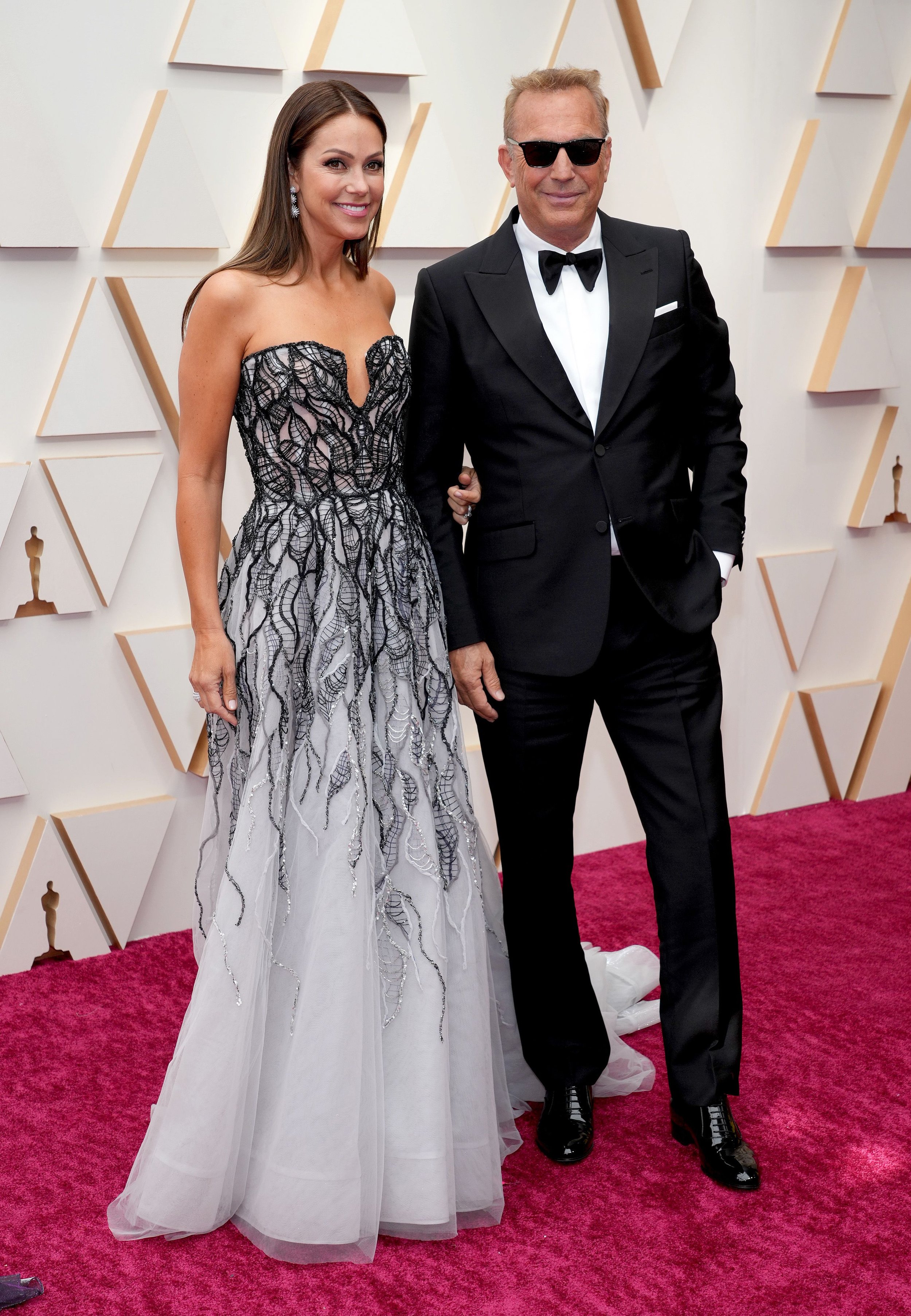 Kevin Costner and Christine Baumgartner posing on a red carpet with a geometric patterned background wall