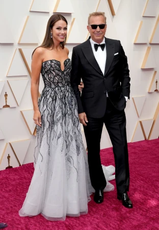 Kevin Costner and Christine Baumgartner posing on a red carpet with a geometric patterned background wall