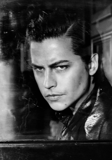 Black and white portrait of a stylish adult male with a slicked-back hairstyle, wearing a shiny jacket, shot through glass with reflections.