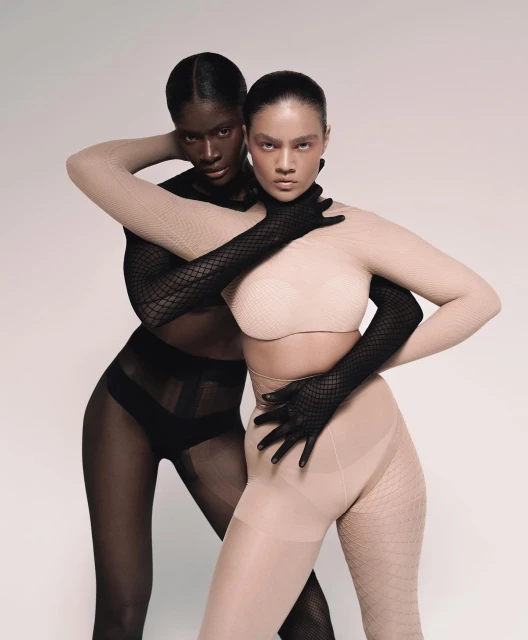Two women with different skin tones posing in coordinated bodysuits and gloves against a neutral backdrop in a fashion setting.